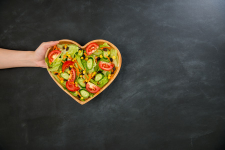 somebody hand showing nutrition vegetable healthy food in wooden heart shaped plate on black chalkboard with high angle view photo.の写真素材