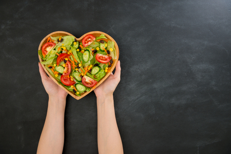 high angle view photo of beautiful hands holding heart shaped wooden plate showing on blackboard with many fresh delicious vegetable.の写真素材