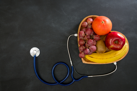 sweet fresh fruit in wooden plate as heart shape with medical device stethoscope isolated on black chalkboard showing healthy care concept.の写真素材