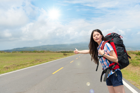elegant female tourist carrying backpack standing on roadside making thumb up gesture looking for car hitchhiking in sunny holiday.の写真素材