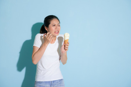 cute attractive female student after eating delicious ice cream feeling sensitive teeth painful uncomfortable and making sadness expression in blue wall background.の写真素材