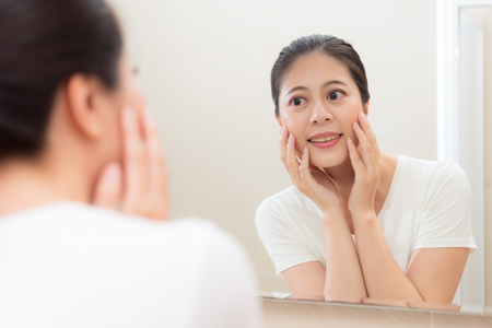 pretty young girl looking at mirror reflection image hands holding on cheek smiling satisfaction face skin healthy situation in bathroom.の写真素材