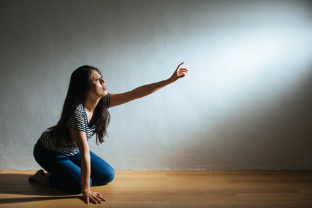 depression lady kneeling on wooden floor ground and reach out hand to light empty area searching helping on white wall background with battered abused woman concept.の写真素材