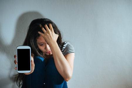 selective focus photo of depressed sadness woman sitting in dark white wall background room holding head crying and showing empty screen mobile smartphone with battered abused women concept.の写真素材