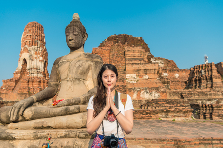 smiling young girl student visiting Ayutthaya famous wat mahathat and standing in front of buddha making praying gesture with face to camera in Thailand travel.の写真素材