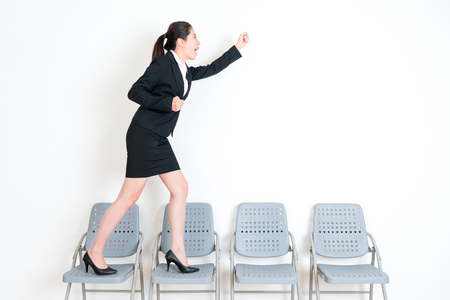 smiling happy fresh graduate standing on chairs and making winner gesture celebrate interview successful in white wall background.の写真素材