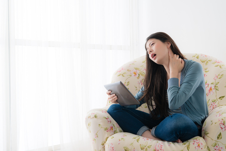 attractive young girl sitting on sofa using mobile pad computer playing game long time getting neck painful and feeling suffered.