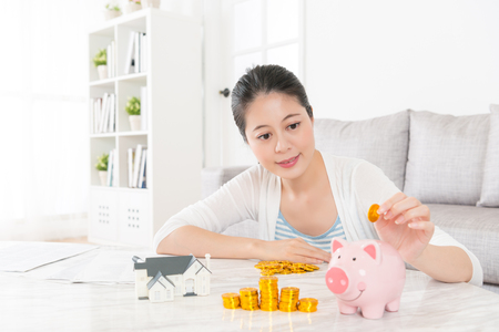 lovely young woman sitting in living room  at home and holding gold coin saving money into pink piggy bank for buying new house with family.の写真素材