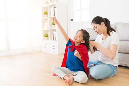 happy cute little kid daughter wearing red cloak play as superhero and making ready to fly posing sitting on living room wooden floor when her mother helping tied hair.