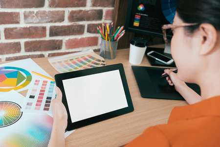 back view of professional young woman designer using mobile digital tablet pad working with blank screen and designing graphic product. selective focus photo.の写真素材