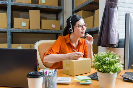 beautiful young online shopping company calling center worker talking with client and looking at computer screen using pen record note on memo paper.の写真素材