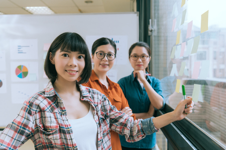 selective focus photo of confident smiling business manager with her working team looking at camera and meeting on glass wall with many sticky paper.の写真素材
