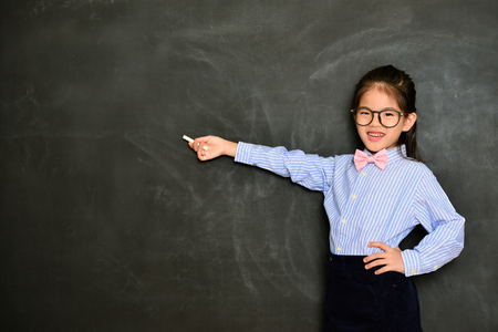 happy confident kid tutor using chalk pointing empty area and showing school studying concept standing in blackboard background.の写真素材