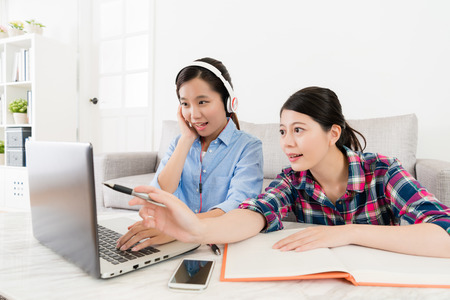 smiling elegant girl student wearing headphone using online e-learning system studying and friend pointing computer screen to assist her finished test.の写真素材