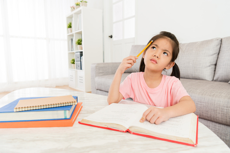 lovely sweet little girl studying school class textbook at home and getting difficulty problem sitting in living room thinking how to solve.の写真素材