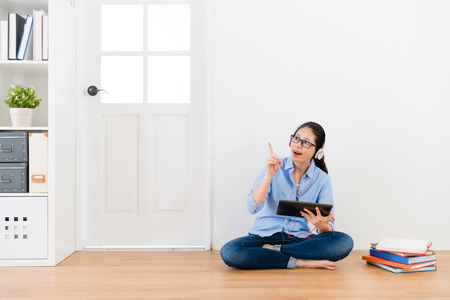 cheerful young female university student having good idea for school homework when she sitting on wooden floor using mobile pad computer listening e-learning education information studying.の写真素材