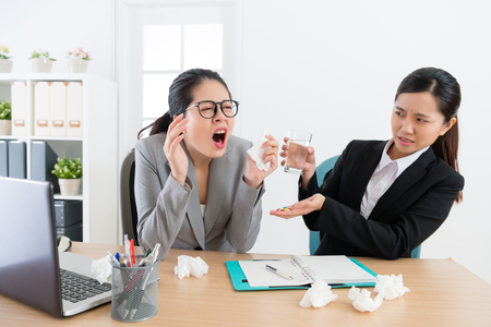 business woman sneezing while her worker partner looking at her feeling fear in office and giving pills with water.の写真素材