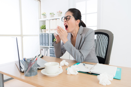 female office worker having nose allergy problem and feeling unwell sneezing in working office during season changing time.の写真素材