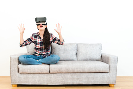 beautiful young woman sitting on wooden floor sofa feeling surprised when she using virtual reality goggles watching 3D simulation video in white background.の写真素材