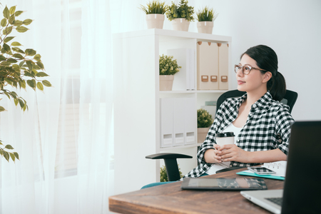 happy beautiful soho business woman holding coffee cup looking at window daydreaming when she working at home in workspace.の写真素材