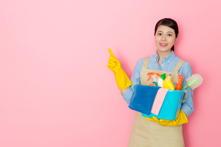 elegant professional housework service woman pointing empty area face to camera and holding many cleaning tool standing in pink background.の写真素材