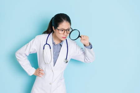 confident pretty female doctor standing in blue background looking down and using magnifier searching health care information.の写真素材