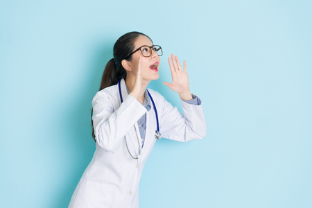 pretty attractive doctor woman using hands as loudspeaker looking at above empty area shouting isolated on blue background.の写真素材