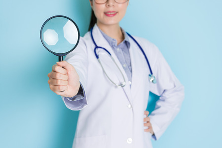 smiling young female doctor standing in blue background and face to camera showing magnifier tool.の写真素材