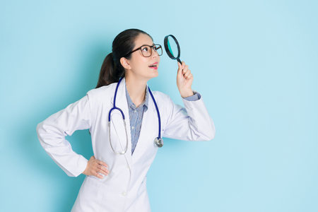 smiling beauty girl doctor carrying stethoscope standing in blue background and using magnifier tool looking at above searching medical information.の写真素材