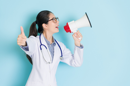 happy attractive woman doctor using loudspeaker announcing medical information and standing in blue background showing thumb up gesture to camera.の写真素材