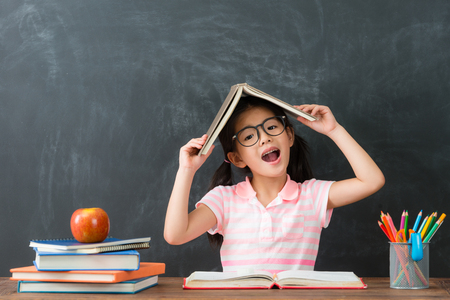 young happy girl children back to school studying sitting in blackboard background and put textbook on head looking at camera.の写真素材