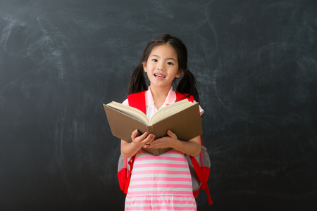 happy beautiful female kid student back to school studying and holding textbook standing in chalkboard background looking at camera.の写真素材