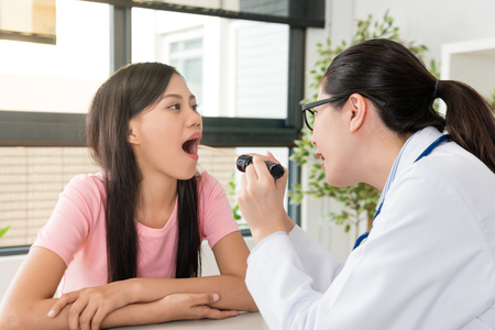 pretty attractive girl opening mouth when clinic female doctor holding electric flashlight to examination her throat status.の写真素材