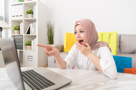 cheerful pretty muslim woman pointing mobile computer screen feeling shocked when she finding online shopping website having super sale.の写真素材