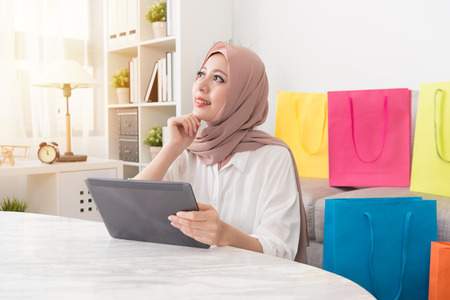 pretty attractive muslim woman looking at air daydreaming thinking when she using mobile pad computer online shopping at home.の写真素材