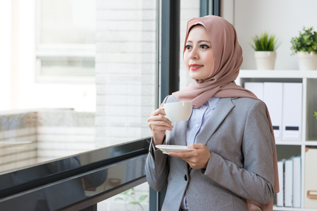 attractive beauty lady muslim business worker holding coffee mug standing in office and looking at window outside daydreaming relaxing.の写真素材