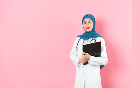 elegant pretty female muslim doctor holding patient record clipboard standing in pink background and looking at camera smiling.の写真素材