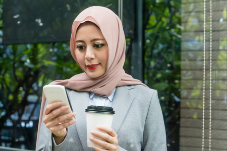 beautiful young muslim businesswoman using mobile smartphone viewing online information when she holding coffee cup standing in outside taking break with window background.の写真素材