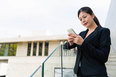 Attractive business woman texting message to her boss with a friendly smile standing outdoors in a black suit.の写真素材