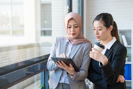 Muslim businesswoman who wears the traditional scarf is telling the co-worker her opinion. They both wearing formal suit standing in the office. One using a tablet, another holding a cup of coffee.の写真素材