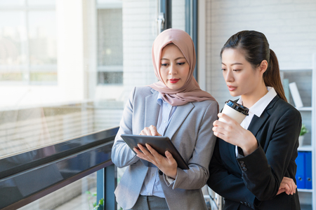 Muslim businesswoman who wears the traditional scarf is telling the co-worker her opinion. They both wearing formal suit standing in the office. One using a tablet, another holding a cup of coffee.