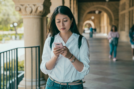 young beautiful student holding her phone and texting her friends for where they are heading to.