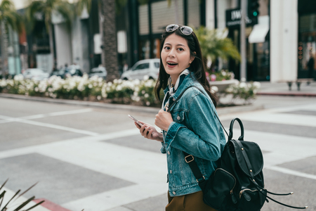 fashion lady cheerfully walking on the road,holding her smartphone and carrying a black backpackの写真素材