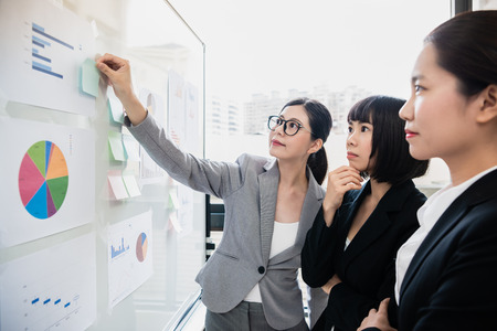 Group of businesswomen in discussion in front of whiteboard at office, manager is being the leader teaching others, everyone is focusing on the note.の写真素材