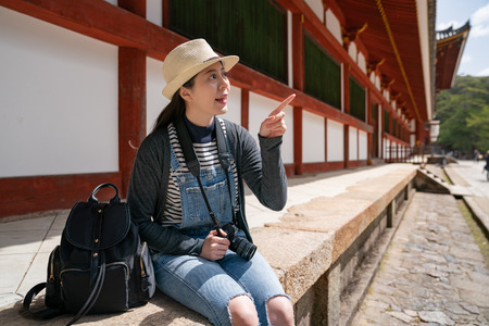 a pretty female photographer relaxing sitting down with her backpack beside her and pointing to the amazing building of the templeの写真素材