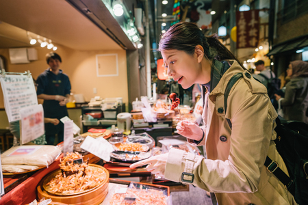 lovely female tourist standing in front of the vendor in the market and pointing to the Japanese street food. the shop owner is smiling at her in the background. Joyful backpacker travel in Japan.の写真素材