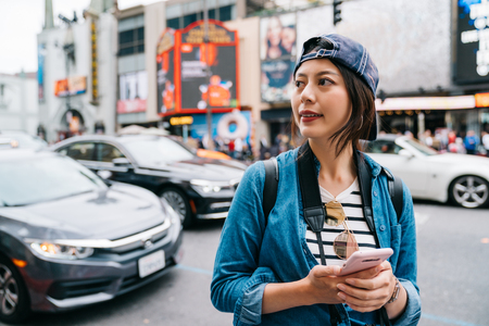 female tourist finding the shop which is having a big sale by her map in the smartphone. Young girl using taxi app on mobile. Asian lady in cap walking in Hollywood street.の写真素材