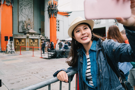 elegant female traveler standing in front of the Chinese Theatre and taking selfie. young girl love taking selfie self-portrait photo on famous sightseeing spot. Joyful tourist travel vacation in US.の写真素材