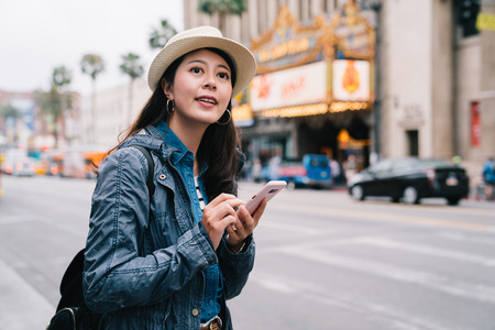 elegant female traveler standing on the road and searching by her cellphone. Young joyful woman expressing positivity, using online map. Spring vacation with bags.の写真素材