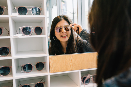 attractive woman trying sunglasses in the glasses shop and looking in the mirror. Asian lady shopping lifestyle. beautiful girl in suitable sunglasses.の写真素材
